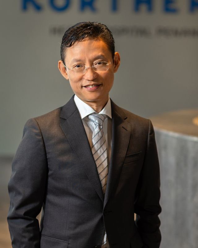 Dr. Tan Chiang Soo, dokter jantung Penang, berdiri di lobi Northern Heart Hospital dengan jas abu-abu tua